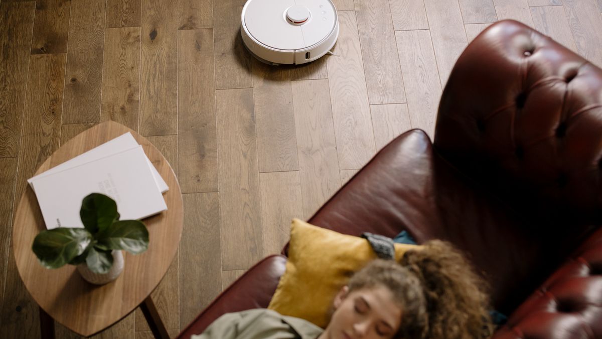 Robot vacuum cleaning a hardwood floor in a living room while a person relaxes on a sofa